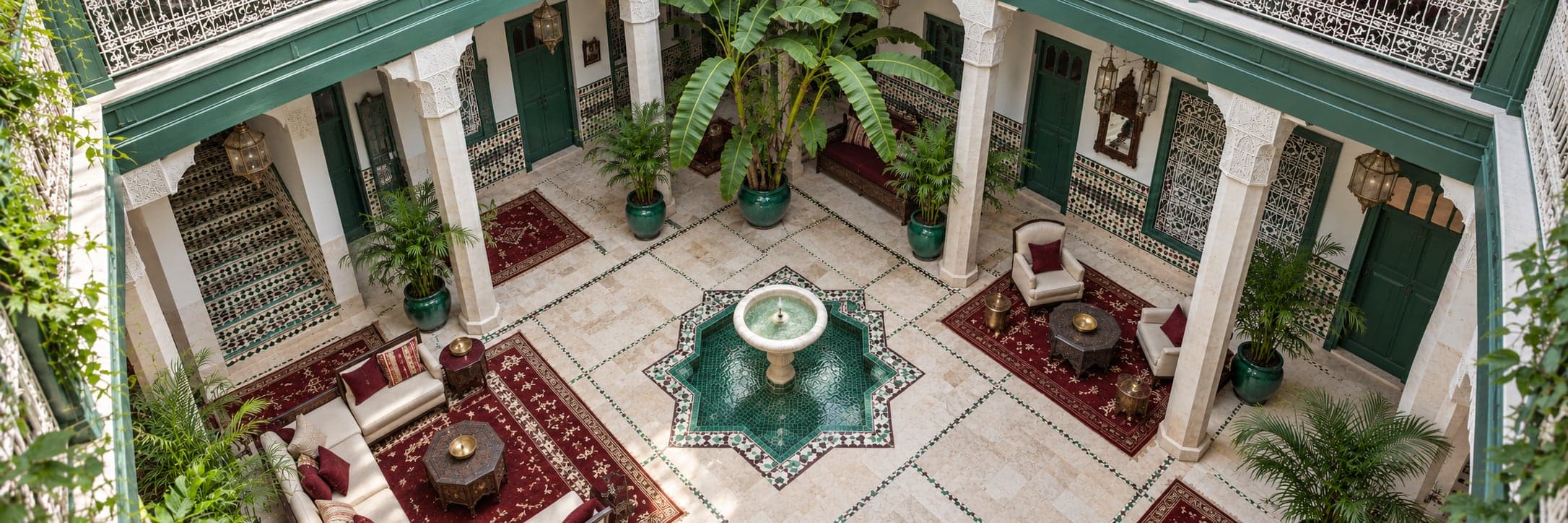 A Moroccan riad courtyard seen from above: a star-shaped green zellige fountain, carved white columns, green doors, potted palms and red rugs with lounge seating
