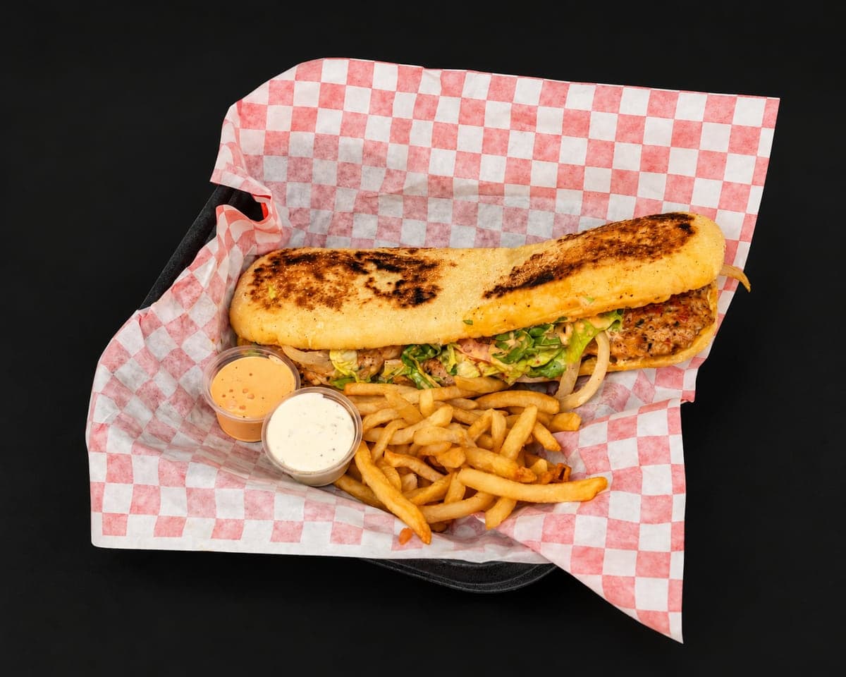 Chicken kefta sandwich in grill-marked batbout with greens and onions, with fries and two dipping sauces in a checkered paper basket on a matte black background