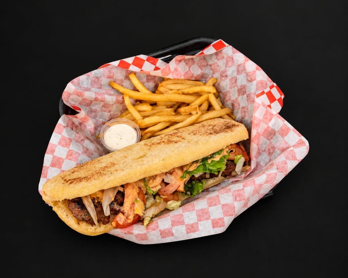 Beef kefta sandwich in golden batbout with tomatoes, onions and greens, with fries and two dipping sauces in a checkered paper basket on a matte black background
