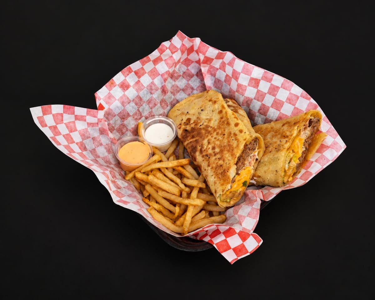 Msemen beef wrap cut in half showing beef and melted cheese, with fries and two dipping sauces in a checkered paper basket on a matte black background