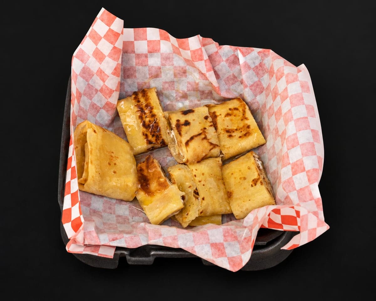 Golden griddled msemen pieces filled with cheese and Nutella in a checkered paper basket on a matte black background
