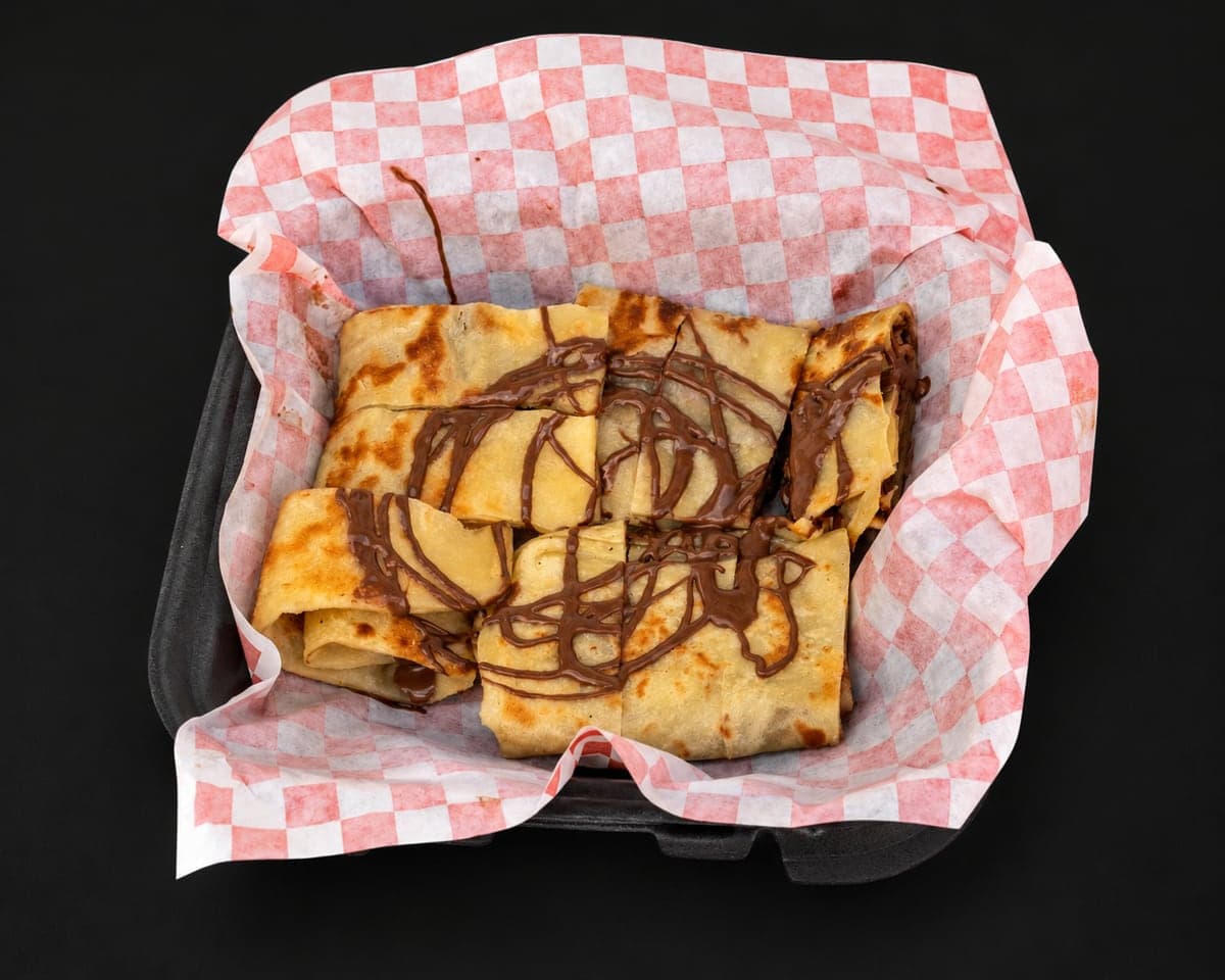 Griddled msemen squares drizzled with Nutella in a red-and-white checkered paper basket on a matte black background